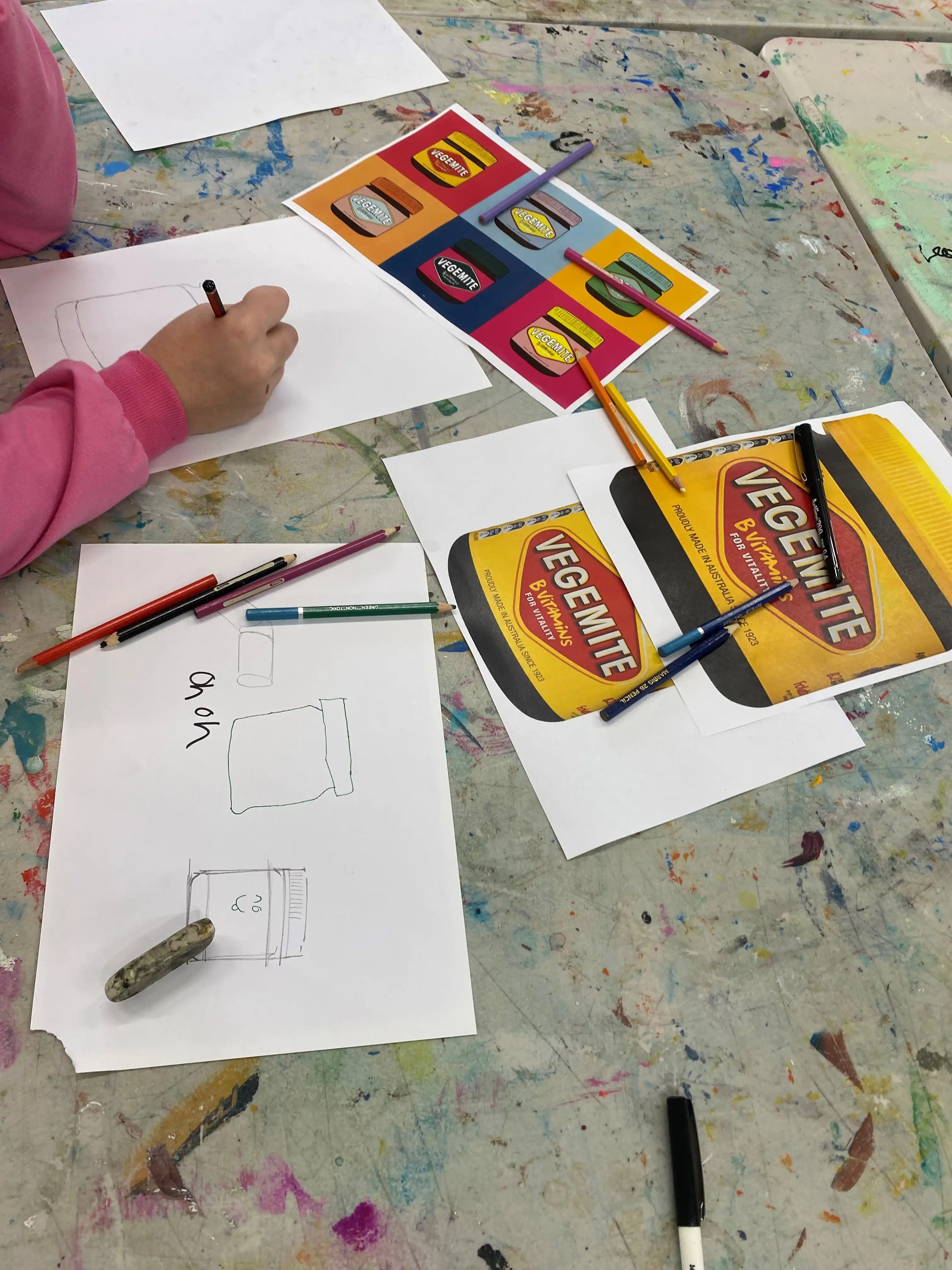 Close-up of a child sketching Vegemite jar designs with printed reference images and coloured pencils on a messy art table.